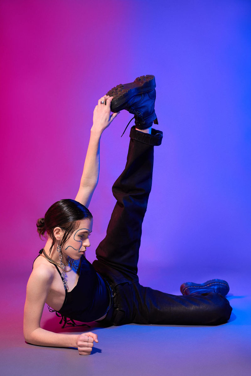 Künstlerisches Portraitfoto in liegender Pose mit pink-blauer Neon-Beleuchtung, schwarzes Styling mit künstlerischem Face-Paint