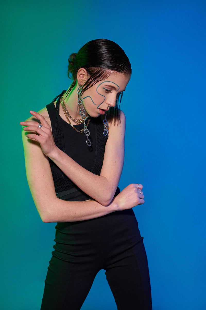 Ganzkörper-Portrait mit dynamischer Armbewegung, grün-blaue Farbbeleuchtung, schwarzes Outfit mit künstlerischem Make-up