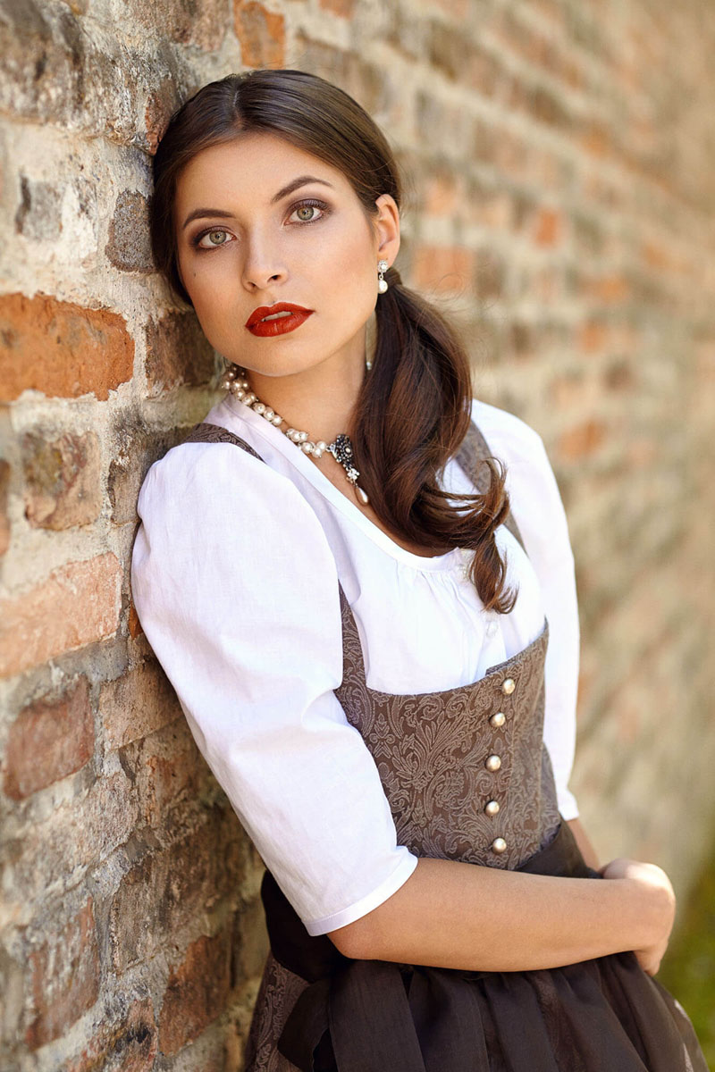 Dirndl Portrait an alter Steinmauer im Freien, Model in grauem Trachten-Dirndl mit rotem Lippenstift, natürliches Outdoor-Licht