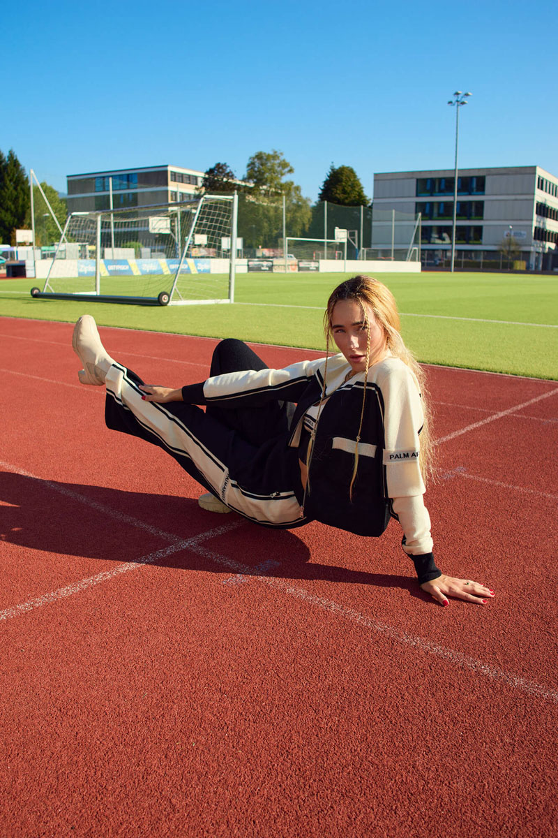 Blonde Frau in schwarz-weißem Palm Angels Trainingsanzug auf roter Laufbahn vor Fußballplatz