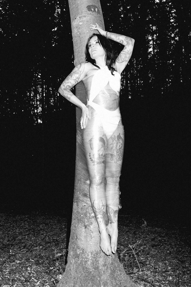 Konzeptuelle Porträtfotografie Traunstein - Künstlerische Aktfotografie Bayern Künstlerische Schwarz-Weiß-Fotografie einer tätowierten Frau in transparenter Folie am Baumstamm im Wald