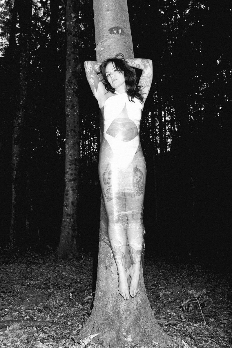 Tätowierte Frau in Frischhaltefolie am Baum - künstlerische Schwarz-Weiß-Porträtfotografie mit verschränkten Armen