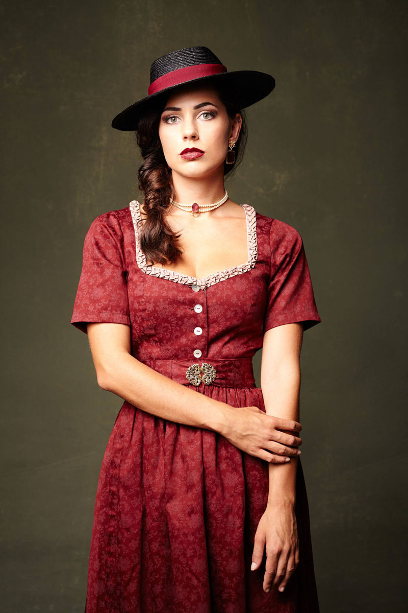 Elegante Frau in rotem Samtdirndl mit schwarzem Hut und Perlenkette - Trachtenfotografie Traunstein